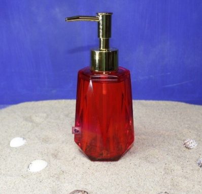 Roter Seifenspender mit goldfarbener Pumpe auf sandigem Untergrund und Muscheln.