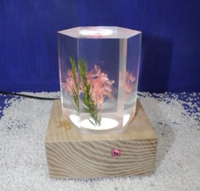Transparente, sechseckige Skulptur mit rosa Blüten und Holzsockel.
